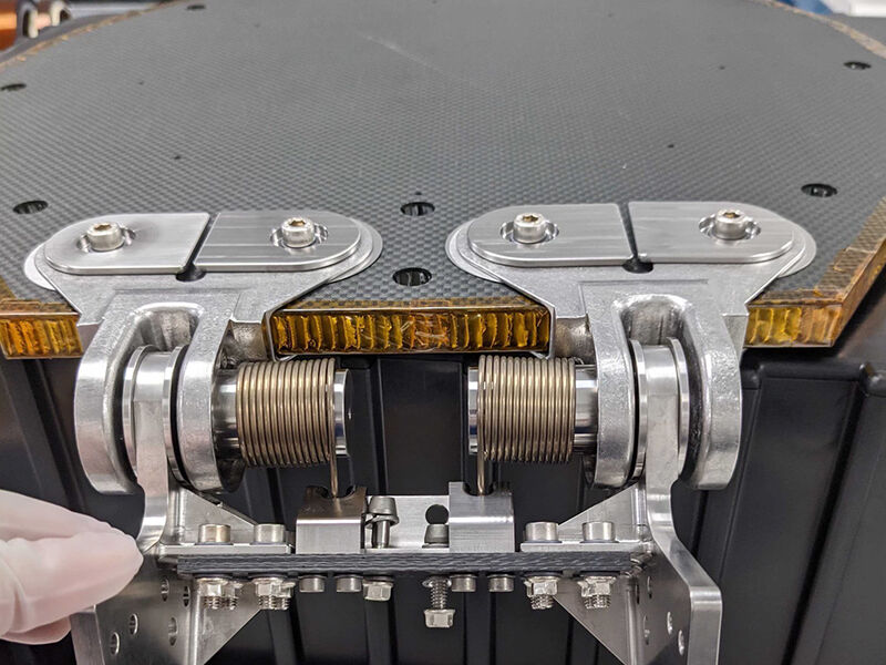 Close up photo of the Psyche cover deployment mechanism attached to the optics enclosure. The mechanism is a metallic, spring-loaded joint. The enclosure cover is a carbon fiber sandwich panel with an aluminum core with kapton edging.  
